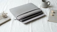 Gray fabric laptop sleeve case with zipper containing multiple laptops and tablets on white desk with coffee mug and notebooks for office organization.