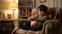 Young boy with dark hair, Caucasian ethnicity, sitting in upholstered armchair holding piggy bank, warm home interior with lamp and bookshelf, financial education concept.
