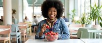Young woman with afro hairstyle and dark skin tone eating fresh strawberries in bright modern office or cafe setting, healthy snacking and wellness lifestyle.