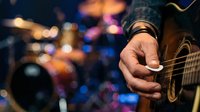 Close-up of male musician's hand playing acoustic guitar during live performance with colorful stage lights and blurred band members in background for music event promotion.