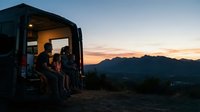 Silhouetted figures sit in an open van door at sunset, gazing at mountain landscape during golden hour. Suitable for travel, adventure, lifestyle, and tourism marketing materials.