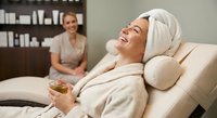 Young woman relaxing in spa chair with towel on head holding herbal tea while therapist sits in background. Wellness and self-care concept.
