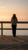 Woman with long dark hair, wearing gray sweater and black pants, standing alone on wooden pier at sunrise gazing toward calm water and distant birds. Serene contemplative moment capturing solitude and peaceful reflection.
