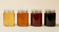 Four glass jars filled with honey in different shades from light golden to dark amber, showcasing natural honey varieties and colors for culinary use.