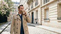 Middle-aged Caucasian businessman in beige trench coat talking on smartphone while walking on cobblestone street in European city architecture.