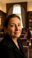 Professional mature Caucasian businesswoman in dark suit smiling confidently in elegant office interior with warm lighting and classic furniture.