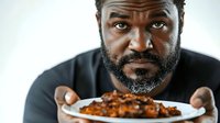 Middle-aged African American male with full beard wearing black shirt, presenting plate of cooked chickpeas or legumes against white background. Suitable for food, nutrition, cooking, and health-related content.