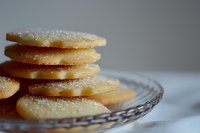 Sugar cookies stacked on vintage glass plate with sparkling sugar crystals, soft natural lighting creates delicate shadows on textured linen tablecloth.