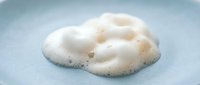 White creamy foam with bubbles on a light blue surface, close-up texture of soap lather or skincare cleanser product.