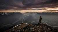 Hiker standing on mountain peak overlooking vast alpine valley with winding river and snow-capped peaks at dramatic sunset, adventure and wilderness exploration.
