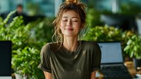 Young woman with messy bun hairstyle relaxing with closed eyes in modern office workspace surrounded by green plants and laptop computer for wellness concept.