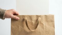 Hand holding brown kraft paper shopping bag with handles against neutral background for eco-friendly retail and sustainable packaging concepts.