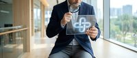 Professional businessman in suit holding digital tablet displaying medical cross symbol in modern office environment with city view background.