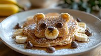 Playful bear-shaped pancake decorated with chocolate chips, banana slices, powdered sugar, and maple syrup on white plate, creative breakfast food styling.