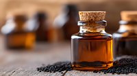 Small glass bottle with amber liquid and cork stopper against scattered black cumin seeds on wooden surface, selective focus on foreground with blurred background.