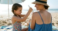 Young girl applying sunscreen to grandmother's back on sunny beach vacation, family skincare protection and bonding moment by ocean.