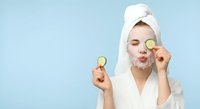 Young woman in white bathrobe and towel holding cucumber slices for natural skincare routine against blue background for beauty and wellness content.