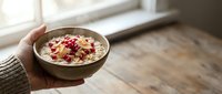 Warm oatmeal bowl topped with dried cranberries and nuts, held by hand near bright window for breakfast, healthy eating, and morning meal content.