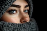 Close up portrait of young Middle Eastern woman with expressive blue eyes and long eyelashes wearing gray knitted winter scarf and hat against dark background.