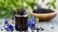 Black seed oil being poured into amber glass bottle with nigella sativa seeds and purple flowers on rustic wooden surface for natural wellness.