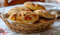 Fresh baked golden bread rolls in woven basket on embroidered tablecloth, crusty homemade pastries with glossy surface and rustic presentation.