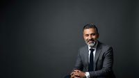 Mature male professional with salt-and-pepper hair and beard wearing dark gray suit and navy tie, smiling confidently with hands clasped, isolated against dark gray background for corporate portraits and business headshots.