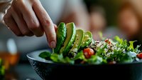 Fresh green salad with sliced avocado being garnished by female hands, close-up view of healthy vegetarian dish with quinoa, tomatoes and mixed greens.