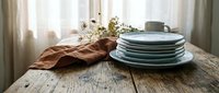 Stacked ceramic plates in muted tones arranged on rustic wooden table with linen fabric and dried flowers for home decor and tableware styling.