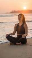 Young Caucasian woman meditating in lotus pose on sandy beach at golden sunset, eyes closed, wearing dark sportswear, ocean waves behind her.