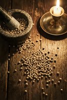 Stone mortar and pestle with coriander seeds scattered on rustic wooden table beside burning candle creating warm atmospheric cooking scene.