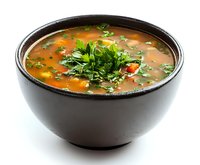 Fresh vegetable soup garnished with parsley in dark ceramic bowl against white background, clear broth with colorful ingredients and aromatic herbs.