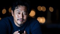 Asian man in dark setting with bokeh lights background, wearing black shirt, looking thoughtfully at camera with gentle smile. Professional portrait capturing authentic emotion.