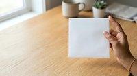 Young adult with medium skin tone holding blank white card or paper at wooden desk with coffee mug and potted plants, suitable for business, marketing, and stationery mockups.