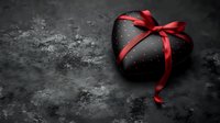 Black heart-shaped gift box with red satin ribbon bow on dark textured surface for romantic occasions, Valentine's Day, anniversary celebrations.