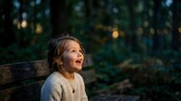 Young girl with dark hair sitting on wooden bench in forest at dusk, looking upward with wonder and contemplation, surrounded by blurred trees and warm bokeh lights.