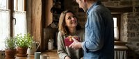 Happy couple enjoying morning coffee together in rustic kitchen with wooden beams and stone walls creating cozy domestic atmosphere.