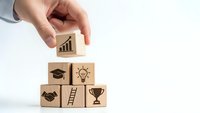 Hand placing wooden cube with growth chart icon on pyramid of business success blocks featuring education, innovation, partnership, career ladder and trophy symbols.