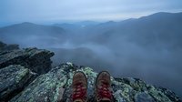 Hiker's boots resting on rocky mountain cliff edge with misty foggy valley and blue hour twilight mountain range in background.