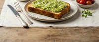 Avocado toast on whole grain bread with microgreens garnish served on white plate with cherry tomatoes and cutlery on rustic wooden table for healthy breakfast.