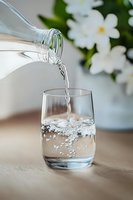 Fresh water pouring from glass bottle into drinking glass with dynamic splash and bubbles, white flowers in soft focus background on wooden surface.