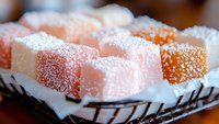 Soft pink and orange Turkish delight cubes dusted with powdered sugar arranged on decorative plate, close-up view showing delicate texture and sweet coating.