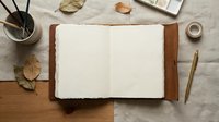 Open blank notebook with leather cover on wooden desk surrounded by autumn leaves, pencils in holder, and coffee cup for creative writing workspace.