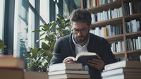 Middle-aged man with beard and glasses reading book in library surrounded by stacked literature and green plants near window.