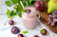 Fresh fruit smoothie in mason jar with pink creamy texture, surrounded by plums, grapes, pear and apple on rustic wooden board, garnished with mint leaves.