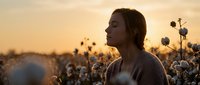 Young woman with closed eyes standing peacefully in cotton field during golden hour sunset, expressing serenity and connection with nature in rural landscape.