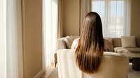 Young woman with long straight glossy brown hair sitting in cozy living room, rear view, soft natural light from window.