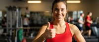 Happy athletic woman giving thumbs up in modern gym after successful workout session with fitness equipment and people exercising in background.