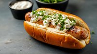 Grilled hot dog in brioche bun topped with fresh herbs and creamy sauce, served with sour cream dip on dark background. Perfect for menu or food advertising.