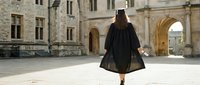 Graduate student walking across university courtyard in black graduation gown and cap, celebrating academic achievement at historic stone campus building.