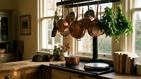 Rustic kitchen interior with copper pots hanging from rack above wooden countertop near sunny window with herbs and cookbooks creating cozy cooking space.